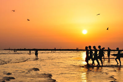 Silhouette people at beach against orange sky during sunset