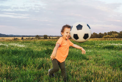 Smiling boy playing with soccer ball on grassy land against sky