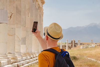 Young male tourist taking picture of old antic ruins with his smartphone on summer day