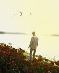 Rear view of man standing by red flowers blooming at lakeshore against clear sky
