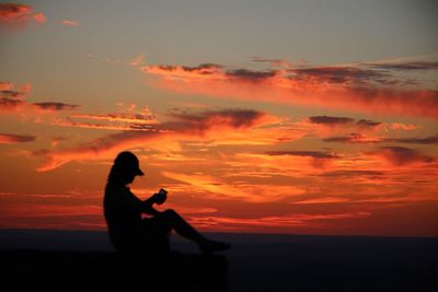Silhouette woman sitting by sea against orange sky