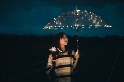 Young woman looking away while standing against sky at night