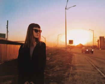 Young woman wearing sunglasses standing by road against sky during sunset