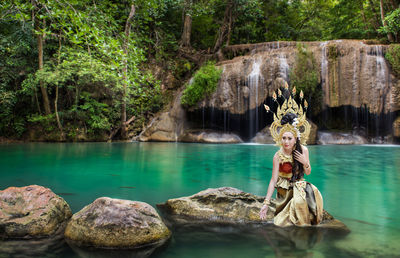 Portrait of beautiful young woman wearing crown and traditional clothing while sitting on rock against lake at forest