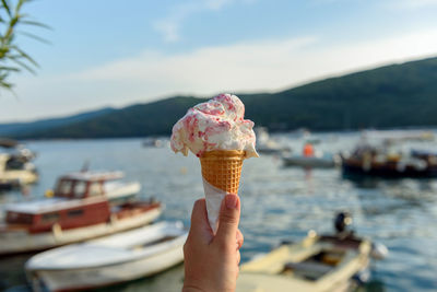 Personal perspective of hand holding two ice cream in cone on waterfront with moored boats in sea