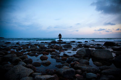 Rear view of man standing on rock at shore against sky during sunset