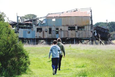Rear view of friends walking on grassy field against abandoned house
