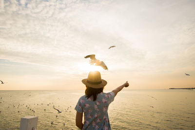 Rear view of woman feeding birds while standing on beach against sky