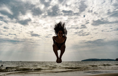Low angle view of young woman jumping at beach against cloudy sky during sunset