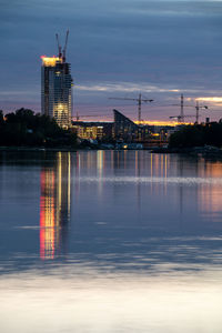 Illuminated factory by river against sky at sunset