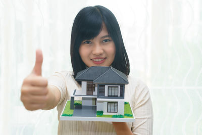 Portrait of smiling young woman holding model house while showing thumbs up