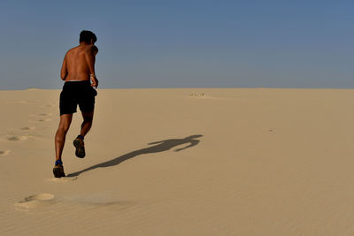 Full length of shirtless young man running on sand at desert during sunny day