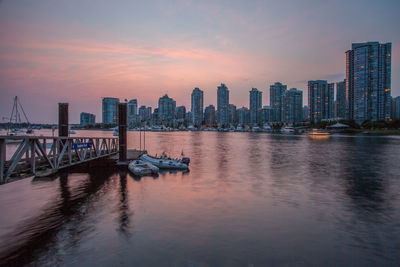 Buildings by lake against sky during sunset in city