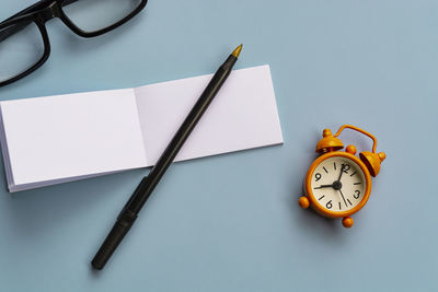 Notepad with reading glasses, pen and alarm clock on blue background. directly above. flat lay.