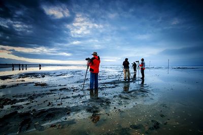 Photographers on wet shore against cloudy sky