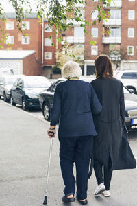 Rear view of elderly woman walking with granddaughter on sidewalk in city