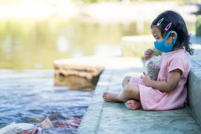 Cute girl wearing protective face mask feeding carp koi fishes at pond.