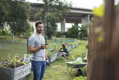 Mid adult man using mobile phone while women working in background at urban garden