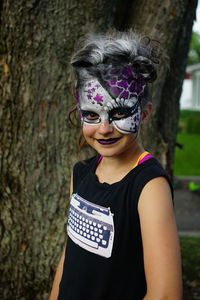 Portrait of smiling girl with face paint standing against tree trunk in park