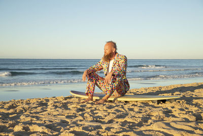 Man wearing colorful suit looking at sea view while sitting on surfboard at beach