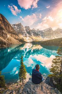 Rear view of young man sitting against lake and mountains on rock during winter