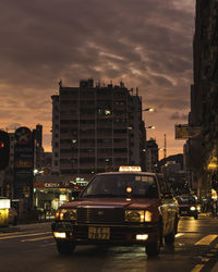 Cars on road by buildings against sky at sunset