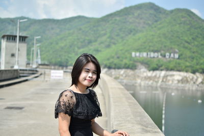 Portrait of young woman standing by retaining wall against sky during sunny day