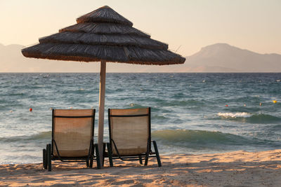 Lounge chairs below thatched roof parasol on shore during sunset
