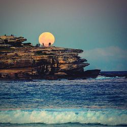 People standing on rock formation by sea against sun at ben buckler