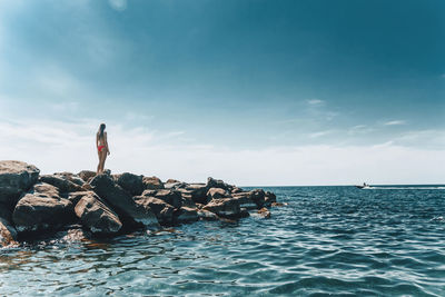 Rear view of bikini woman standing on rocks by sea against sky