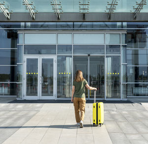 Young woman traveler carrying yellow suitcase next to entrance to airport outside tourism concept 