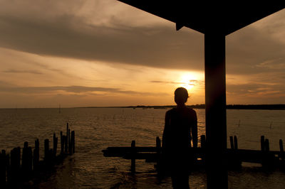 Silhouette woman standing by wooden posts at sea shore during sunset