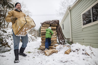 A young child helps his father collect wood from large stack in snow