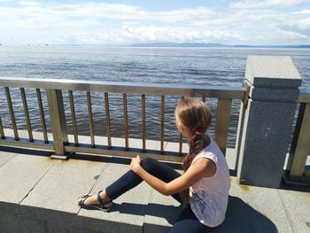 Girl sitting by railing and sea against sky