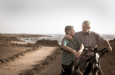 Smiling couple with bicycles standing on land against sea 