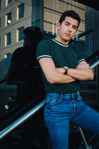 Portrait of young man with arms crossed standing against glass wall