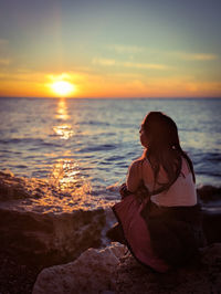 Side view of woman sitting on rock at beach against sky during sunset