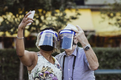 Senior woman taking selfie on smart phone standing by man in park during covid-19