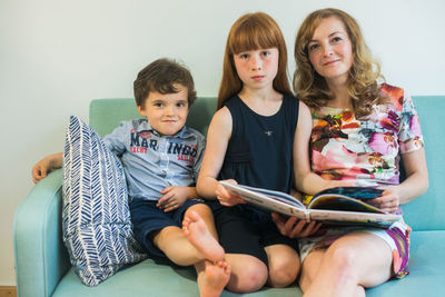 Portrait of mother and children reading book while sitting on sofa at home