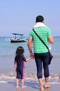 Rear view of father and daughter holding hands while standing at beach against sky