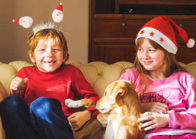 Smiling sibling with dog wearing santa hat sitting on sofa at home