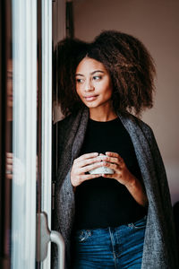 Thoughtful woman drinking coffee while looking through window at home