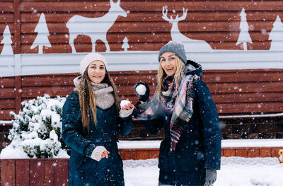Two young happy smiling women in the snowfall throw snowballs at the camera.