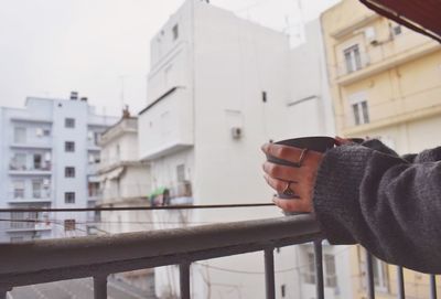 Cropped image of hand holding mug while standing by railing at balcony in city