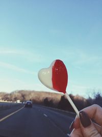 Cropped hand holding heart shape lollipop against sky on road