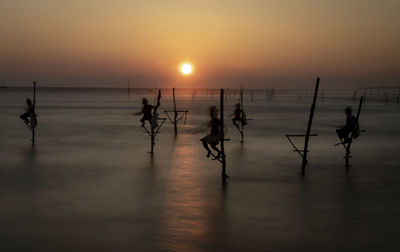 Silhouette people on wooden post at beach against sky during sunset