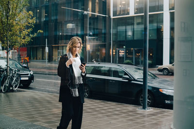 Businesswoman using smart phone while walking on sidewalk against building in city