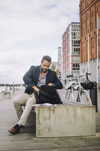 Full length of male entrepreneur searching in backpack while sitting on bench at promenade