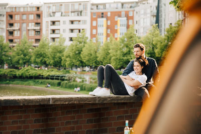 Young man with smiling female partner looking away while sitting on retaining wall