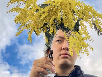 Portrait of young man holding golden wattle flowers in vintage vase against cloudy blue sky.